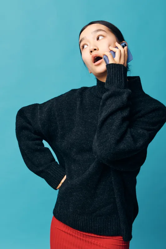 Woman in black sweater and red skirt talking on phone with surprised expression against blue background. Female person communicating and holding smartphone in casual outfit.
