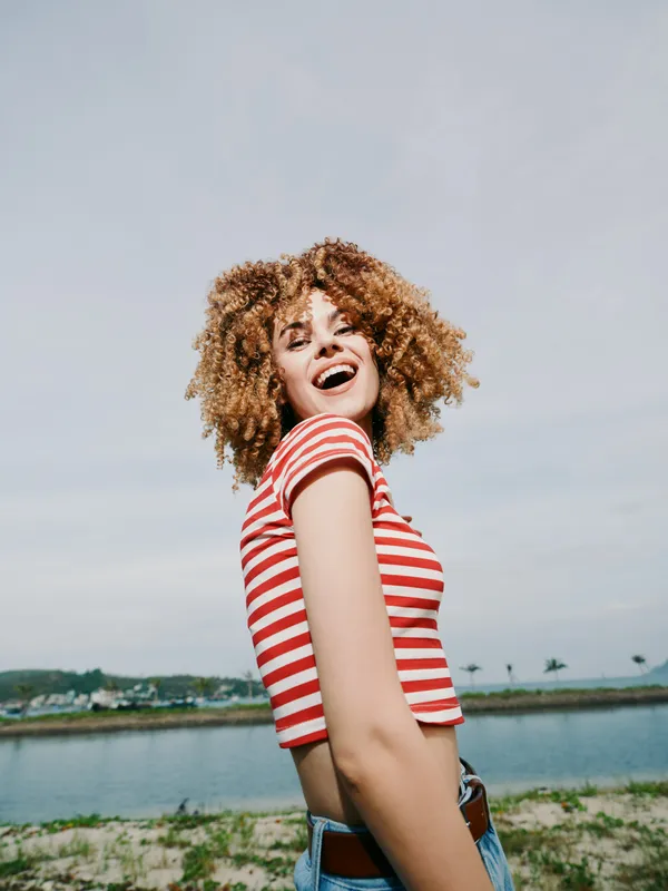 Joyful young woman stands outdoors by a lake, bright smile, curly hair, striped red and white top, casual fashion, sunny day, natural light, candid portrait, lively summer mood.
