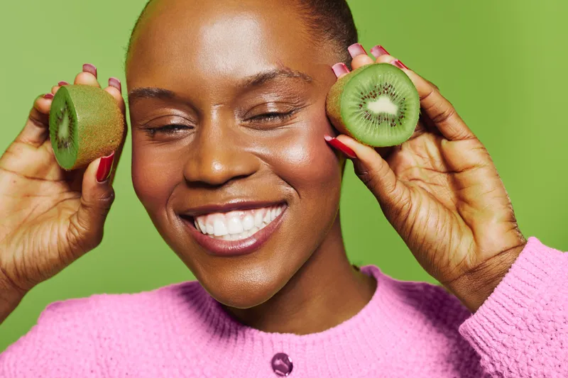 Happy black woman with dark skin smiles holding two halves of fresh kiwi fruit near face. Bright green background and pink sweater create vibrant contrast. Concept of healthy lifestyle and wellness.