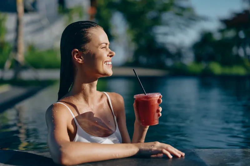 A young woman lounges poolside, sipping a refreshing drink and enjoying a sunlit smile as water sparkles nearby.