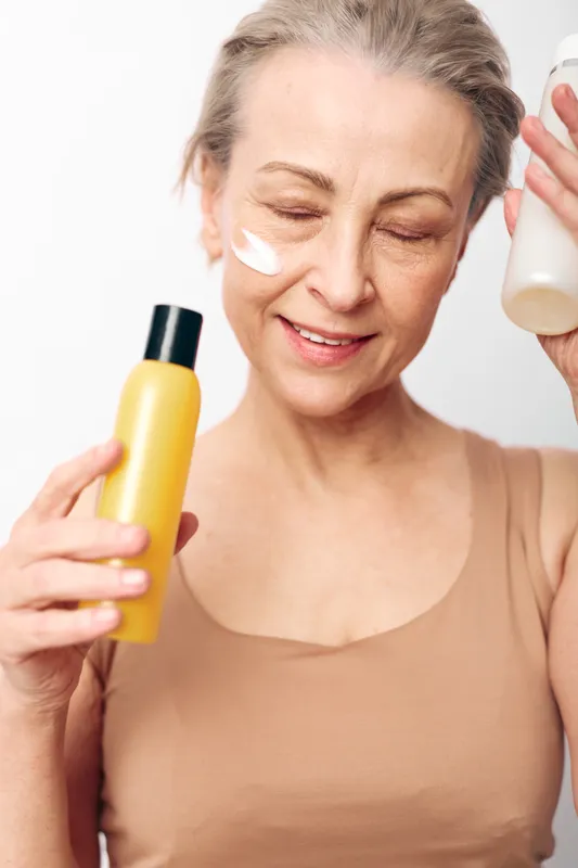 Mature woman with light skin applies cream on cheek while holding two cosmetic bottles. Concept of skincare, aging care, beauty routine, and natural wellness at home.