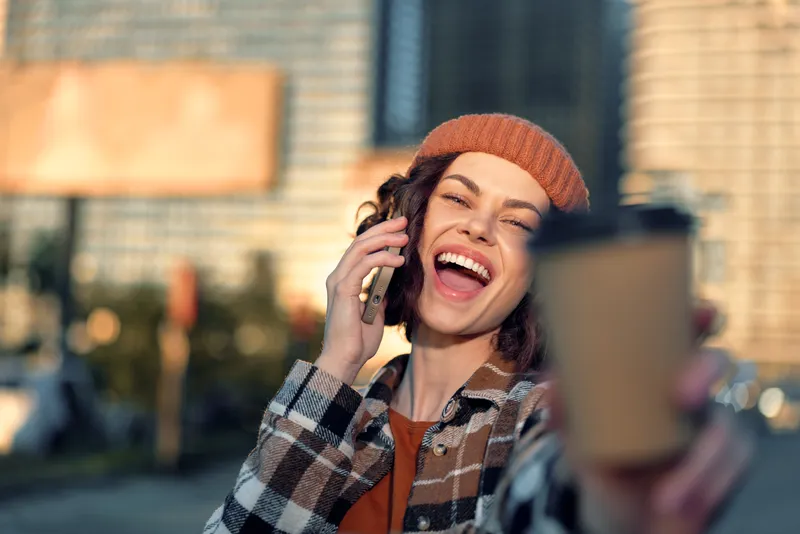 woman coffee phone smile urban candid golden beanie portrait of a joyful young woman talking on mobile and offering a takeaway cup, authenticity, mindful living and emotional storytelling vibe