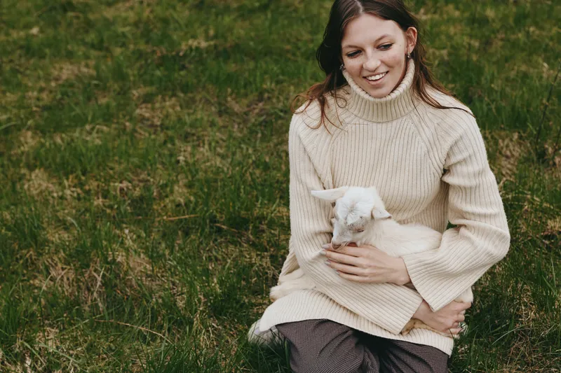 A young woman in a beige sweater sits on grass holding a small white baby goat while smiling gently, capturing a peaceful moment of connection in a natural outdoor setting.