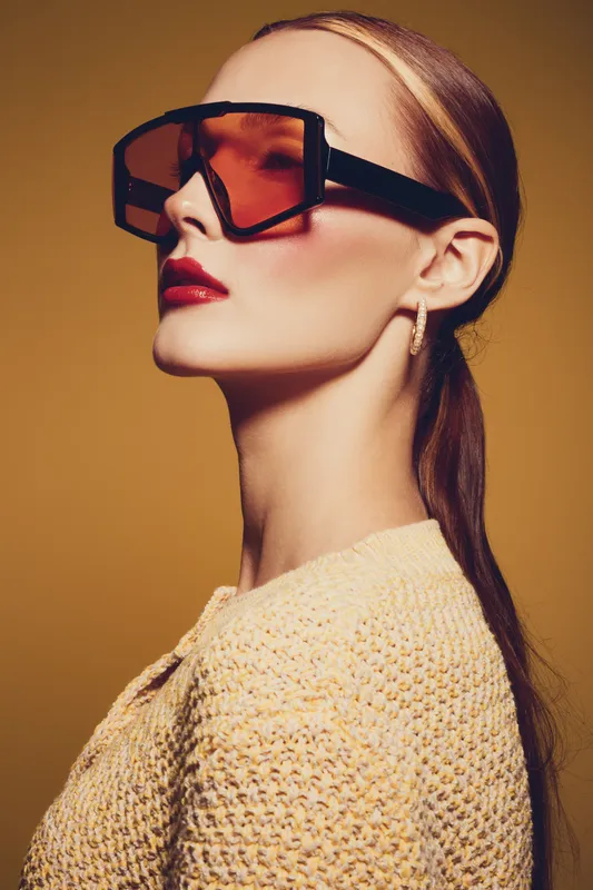 Stylish woman with sleek hair wears oversized tinted sunglasses and red lipstick. She poses confidently against a warm brown background in a textured beige sweater. Fashion concept.