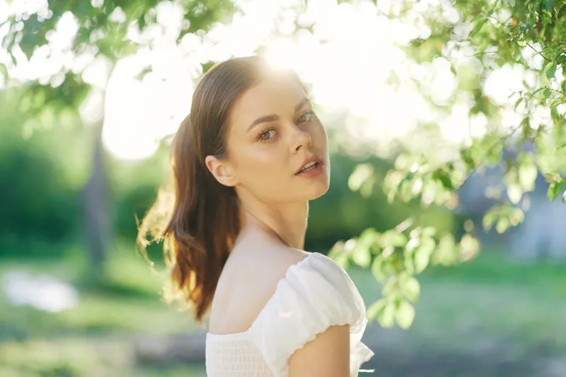 Beautiful young woman with glowing skin and soft smile, surrounded by greenery in a sunny outdoor setting, representing natural beauty and tranquility