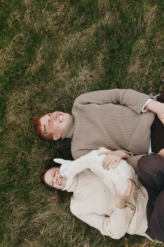 Young couple lies on grass smiling while holding a white baby goat in cozy sweaters during a relaxed outdoor lifestyle moment in autumn.