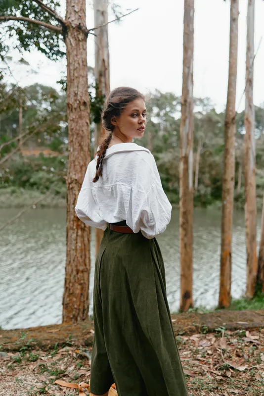 A young woman in a vintage outfit stands by a serene lake surrounded by tall trees, exuding a peaceful and reflective mood