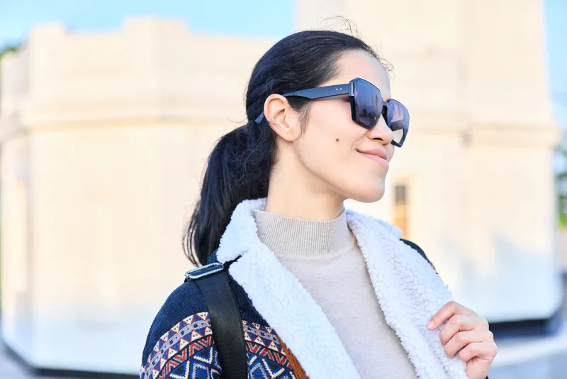 Sunglasses, woman outdoor fashion travel with backpack, stylish outerwear, smiling in sunlight, candid moment of exploration and confidence in a bright outdoor setting