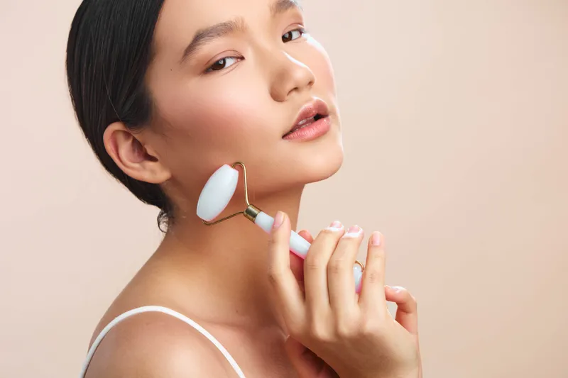 asian woman applies a facial roller in a soft studio setting, showcasing a calm skincare routine with gentle lighting and a smooth complexion, conveying wellness and care.