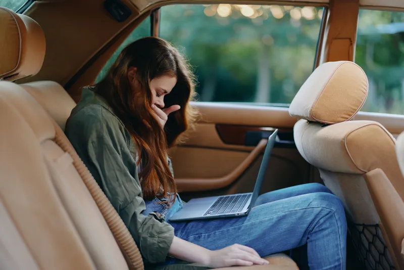 Thoughtful young woman working on a laptop in the backseat of a car, showing concentration, dressed casually in a green jacket and jeans, with nature background.