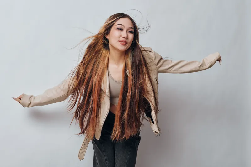 Young woman with long hair wearing jacket dancing with arms outstretched against light background. Happy girl in casual clothes expressing freedom and joy in studio portrait.