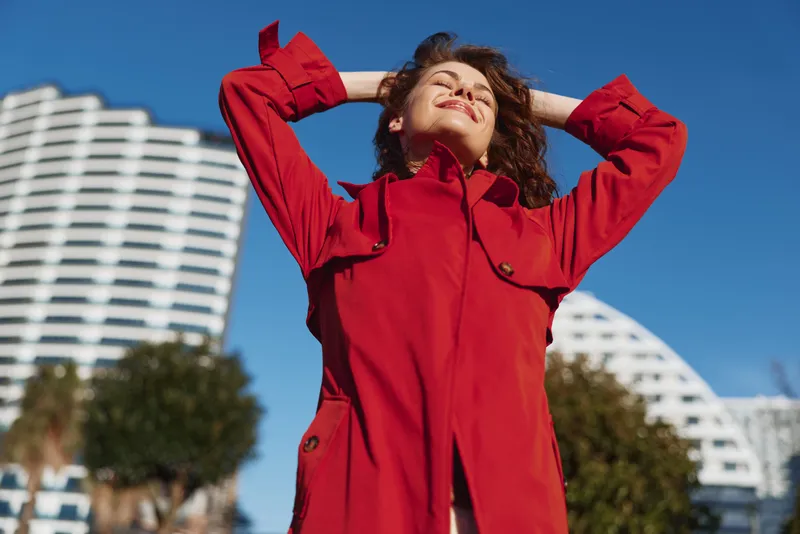 Young woman with curly hair enjoys a sunny day outdoors wearing a bright red coat while stretching with closed eyes and a joyful smile against clear blue sky.