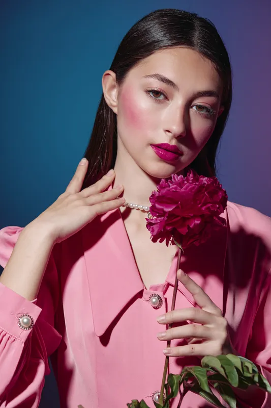 Fashion woman in pink blouse holds dark red flower near face. Creative beauty portrait with elegant pose on dark blue background. Concept of fresh style and softness.