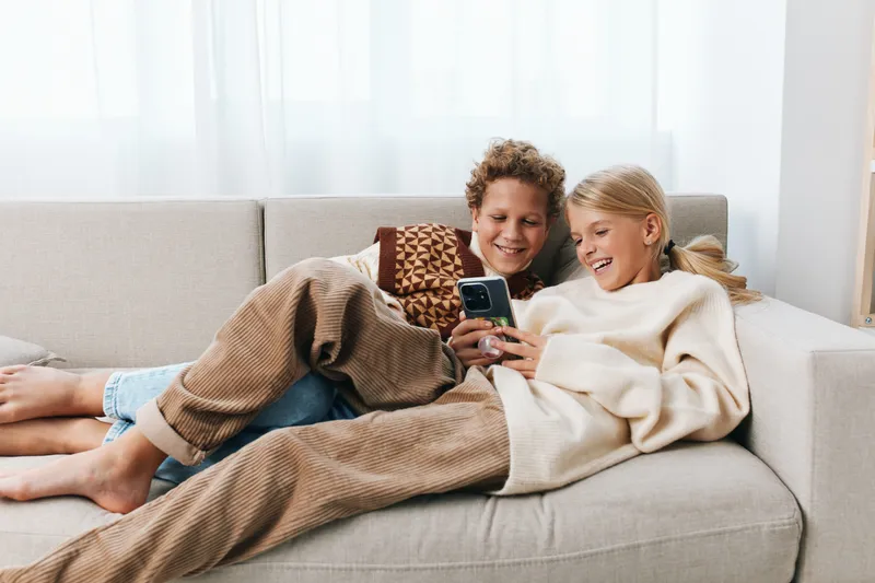 Authentic moment of siblings on a cozy sofa, sharing a smartphone, capturing casual play and daily school life within a warm, relaxed lifestyle setting.