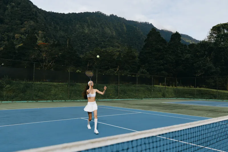 Female tennis player in white sportswear serving on outdoor blue court surrounded by greenery, showcasing determination and focus