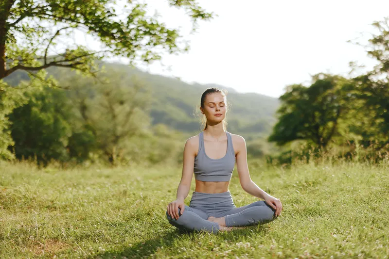 Young woman practicing yoga outdoors in a serene green landscape, wearing a grey sports outfit, embodying tranquility and mindfulness through meditation