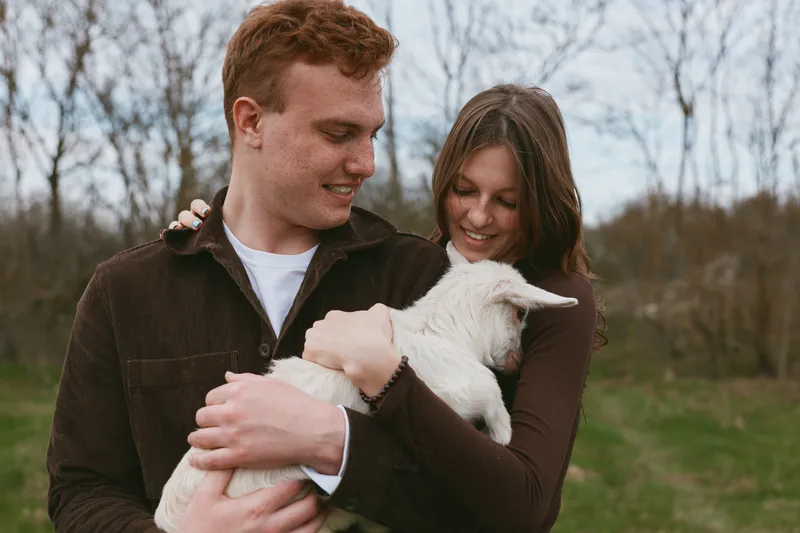A loving couple outdoors cradle a white goat kid, smiling as the gentle animal rests in their arms. A warm, candid portrait of affection, rural life, and animal companionship.