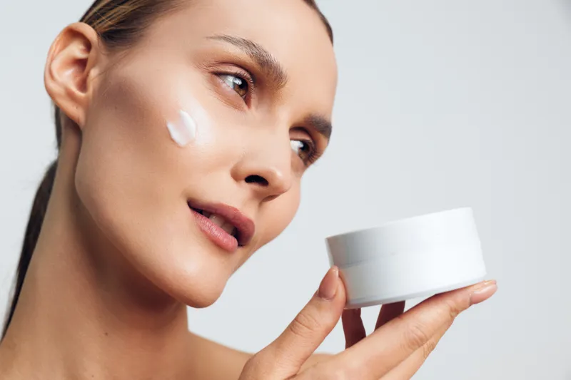 Close-up of a smiling woman with a dab of white facial cream on her cheek, against a clean backdrop. Skincare, wellness, self-care concept. Soft studio lighting creates a warm, inviting mood and glow.