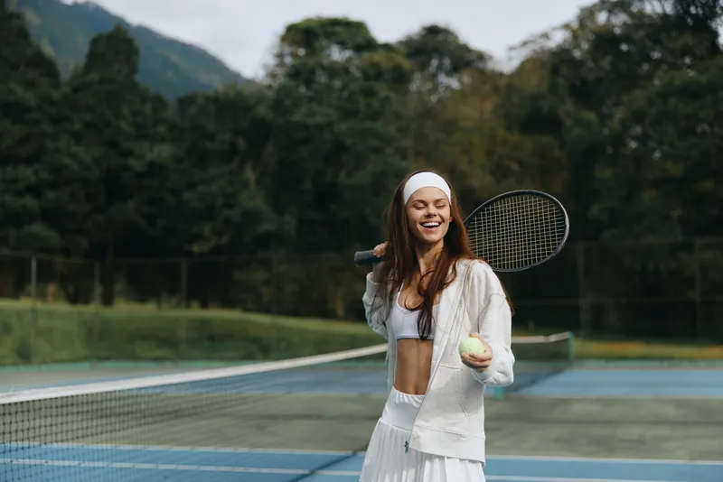 Happy female tennis player in white sportswear holding a tennis ball on a sunny day, conveying joy and energy on a tennis court surrounded by greenery