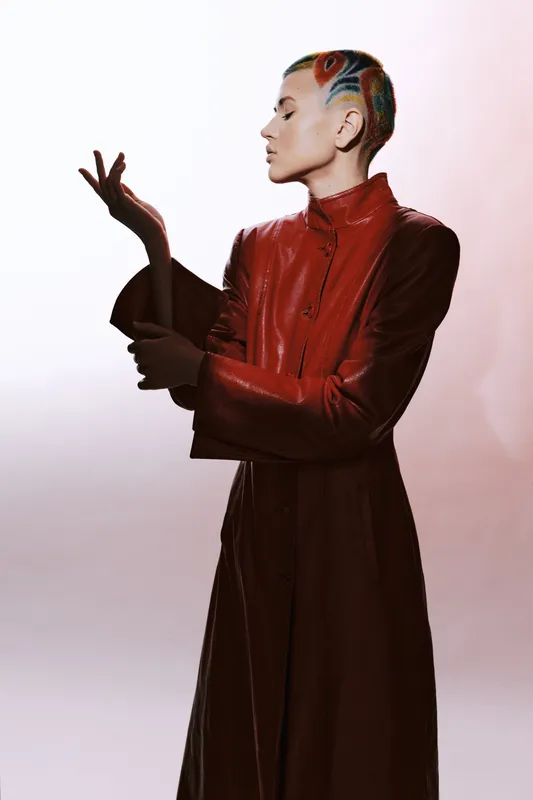 Woman with colorful short hair wearing a red leather coat posing gracefully with a calm expression against a light background.