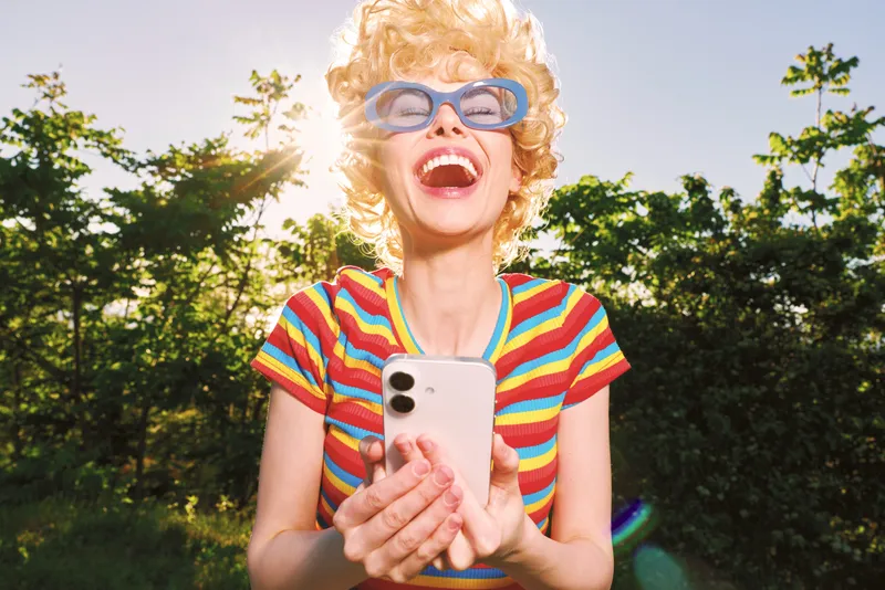 Bright joyful child with blonde hair wears a striped shirt and blue sunglasses, holding a phone, outdoors under sun, smiling widely, playful mood, candid lifestyle shot with warm light and retro vibe