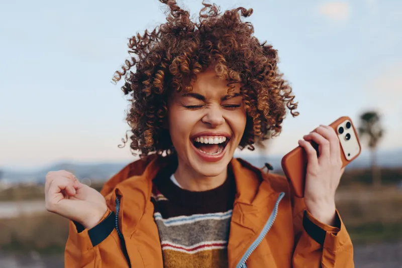 Woman lifestyle smiling with curly hair wearing orange jacket outdoors, laughing while talking on smartphone, warm daylight, casual lifestyle moment, communication and joy today