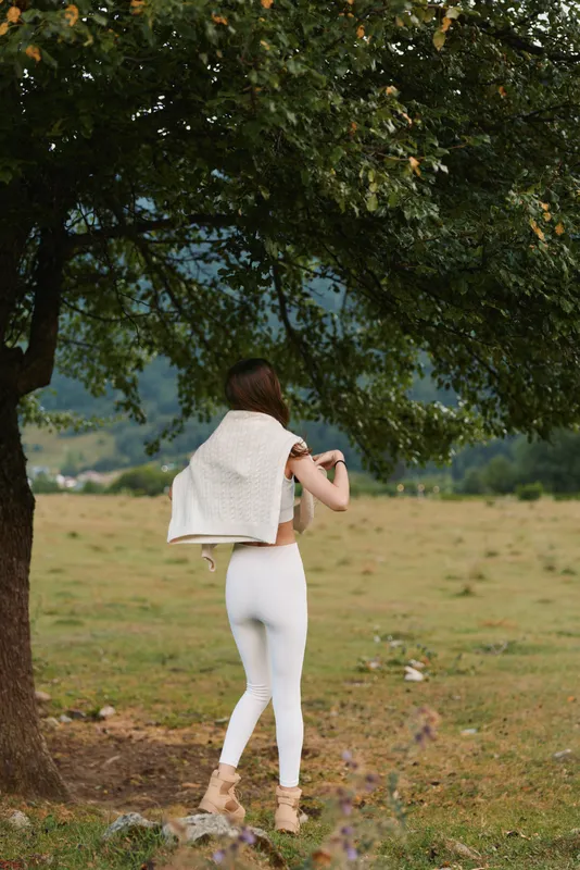 Young woman wearing white athleisure walks under a leafy tree across a sunlit field, savoring a peaceful outdoor moment with a carefree countryside lifestyle.
