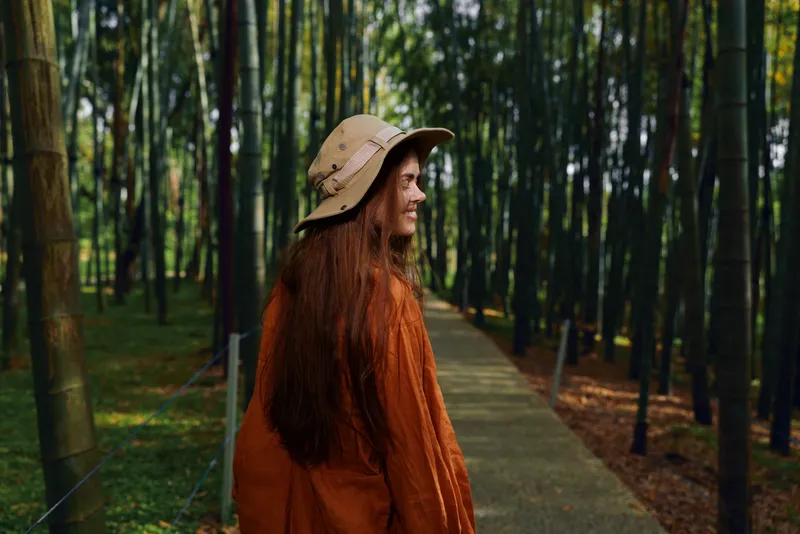 Woman girl with longhair and hat and smile on a path through a bamboo forest, wearing orange coat; portrait of travel and nature, walking on boardwalk in serene outdoor scenery.