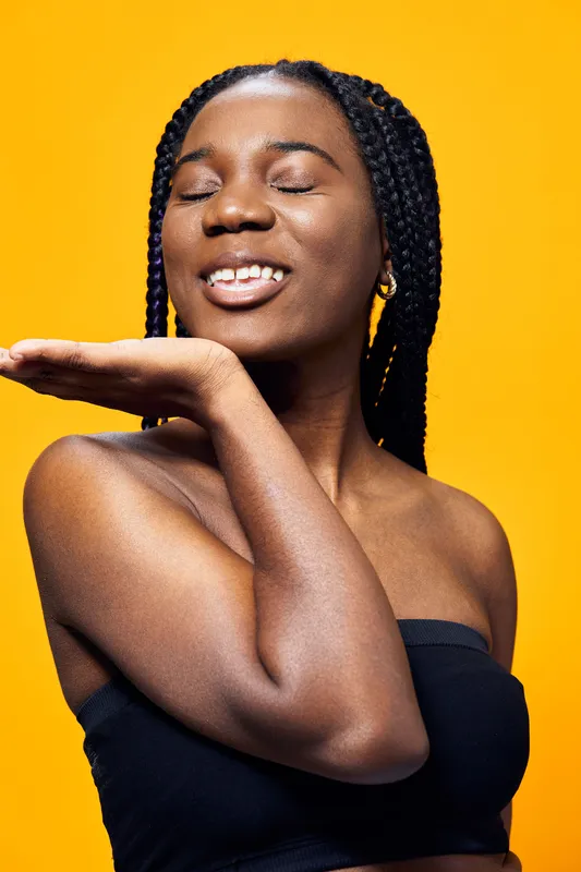 Joyful young woman with braided hair against a bright yellow background, expressing happiness and confidence while wearing a black top