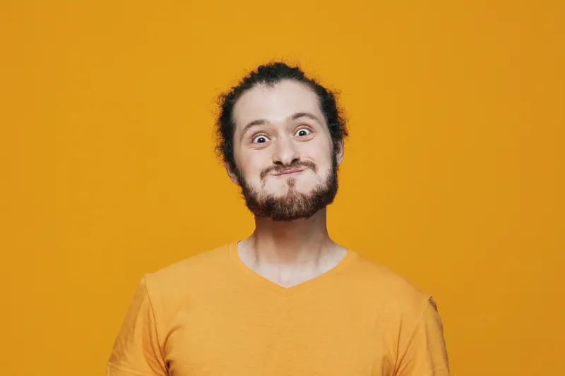 Young man with an expressive face making a funny grimace in a vibrant orange t shirt against a bold yellow background, showcasing joy and playfulness