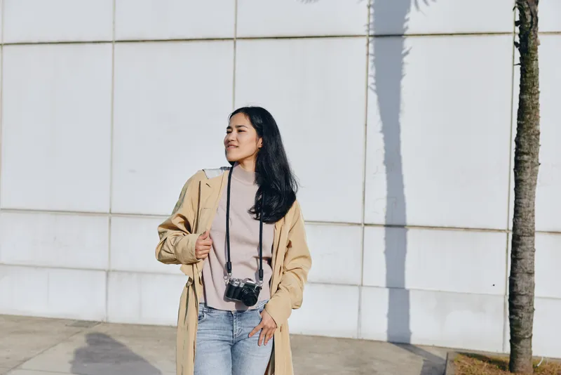 Camera, woman, coat, street, outdoor, fashion, casual, urban, sunlight, pose for lifestyle photography featuring stylish jacket, relaxed stance and candid urban moment