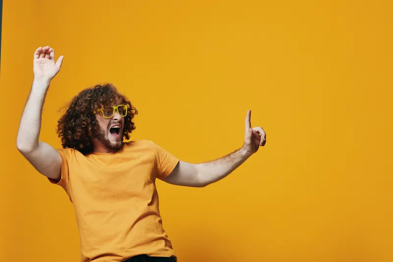 Energetic young man with curly hair wearing sunglasses and yellow shirt, dancing against a vibrant yellow background expressing joy and positivity