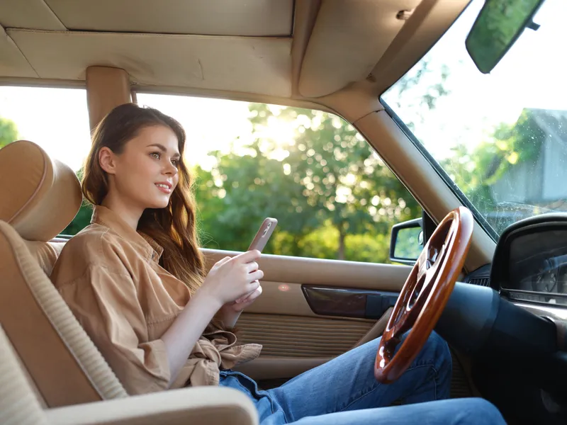 Young woman enjoying a casual drive in a vintage car, using her smartphone while wearing a stylish brown shirt, surrounded by greenery.