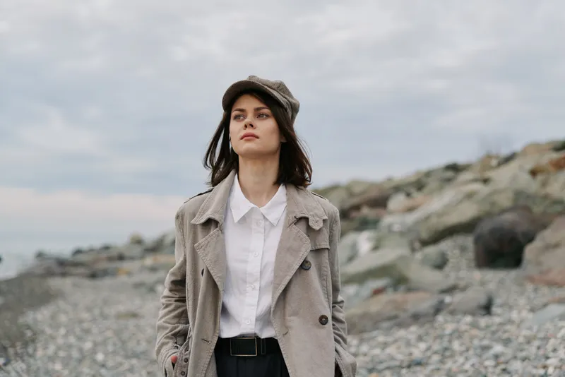Young woman in beige coat and hat stands on rocky beach under cloudy sky. Casual autumn fashion look with confident expression. Natural outdoor setting with muted colors and calm mood.