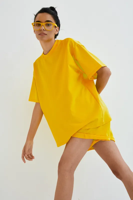 Young woman wearing bright yellow oversized t-shirt and shorts, posing with stylish yellow glasses against plain white background, casual summer outfit.