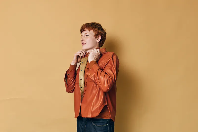 Young man with red hair wearing orange jacket and beige shirt looking thoughtfully sideways against beige background with casual style and confident expression standing.