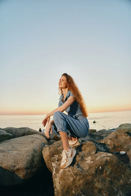 Lifestyle woman in modern boho-western denim street style sitting on rocks near the sea during golden hour with film color mood and relaxed expression.