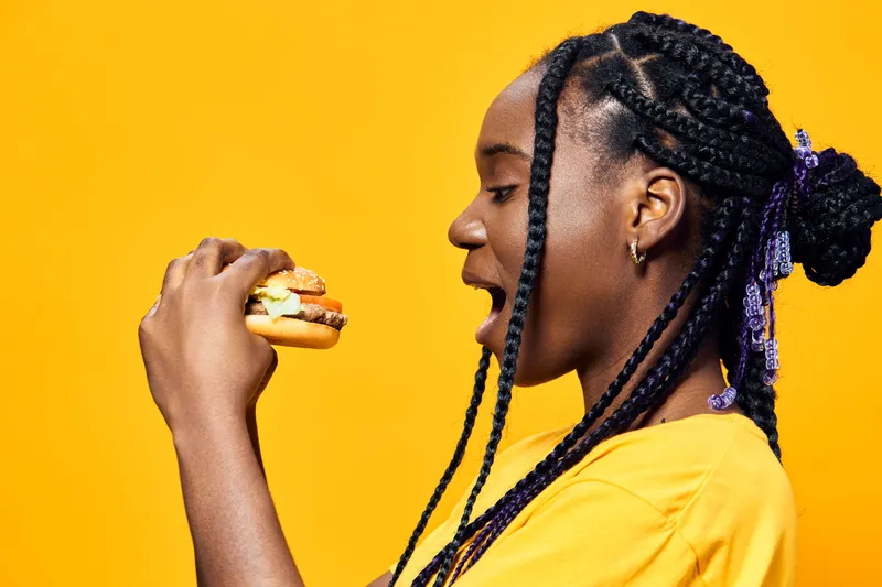 Young woman with braided hair enjoying a colorful burger against a bright yellow background, conveying a sense of joy and healthy eating concepts