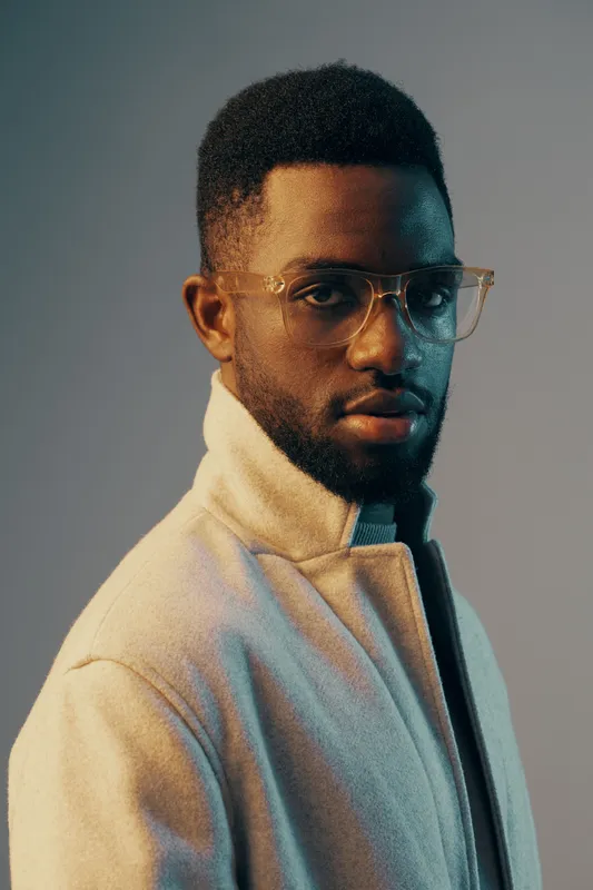 Stylish young African man with glasses in a light colored jacket, set against a gradient background, conveying a modern and confident look