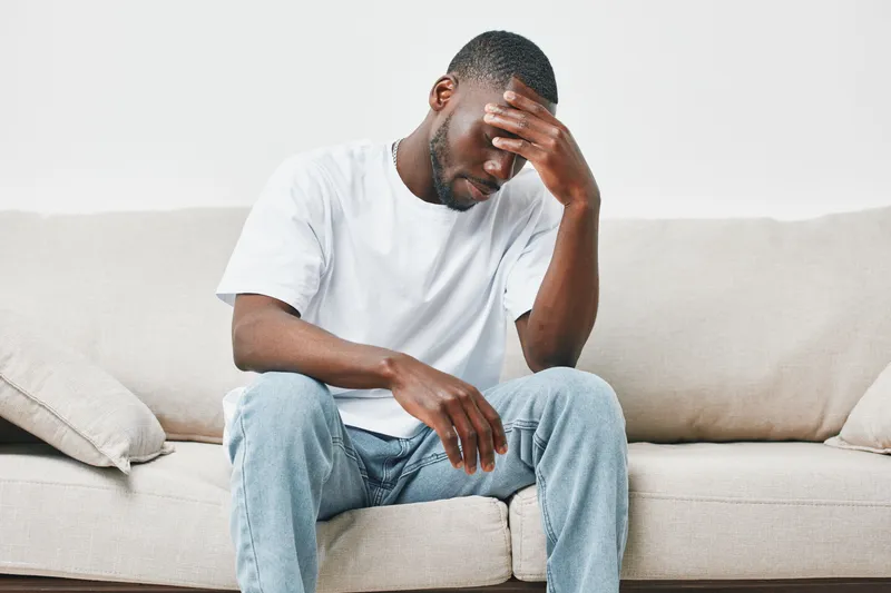 A young African American man sitting on a couch, looking stressed and thoughtful, dressed in a plain white t shirt and blue jeans, reflecting deep emotions