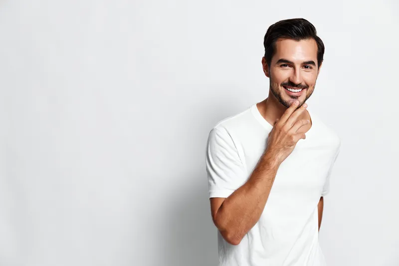 Smiling man wearing a white t-shirt standing confidently with a charming smile, isolated on a plain light background. Person lifestyle concept.