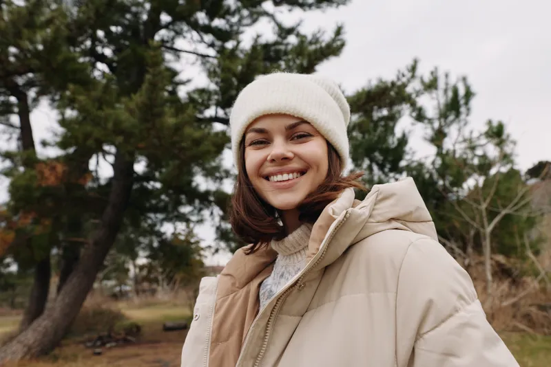 Woman smiling in a warm jacket and knit beanie outdoors in a park, with trees in the background, conveying casual winter fashion and relaxed mood for lifestyle photography.