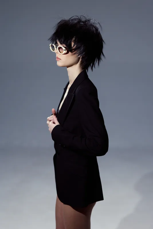 Stylish woman with short hair and sunglasses wearing a black blazer posing on gray background in modern fashion style.
