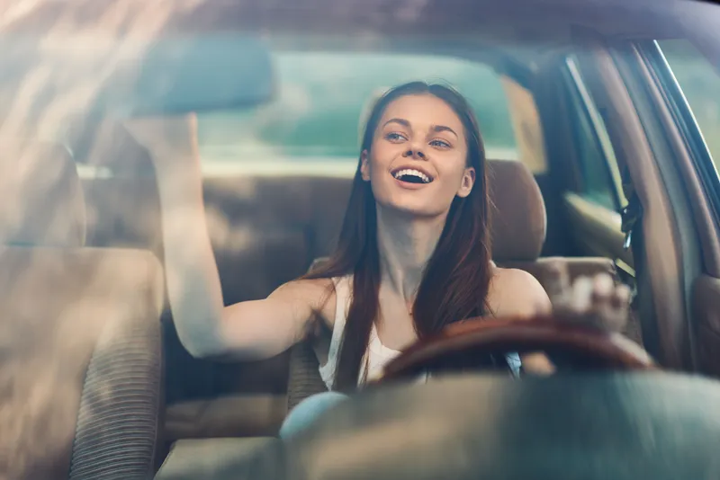 Young woman in a car smiling at the camera, enjoying a sunny day, with vibrant colors and a cheerful ambiance. Joyful expression captures spontaneity and happiness.