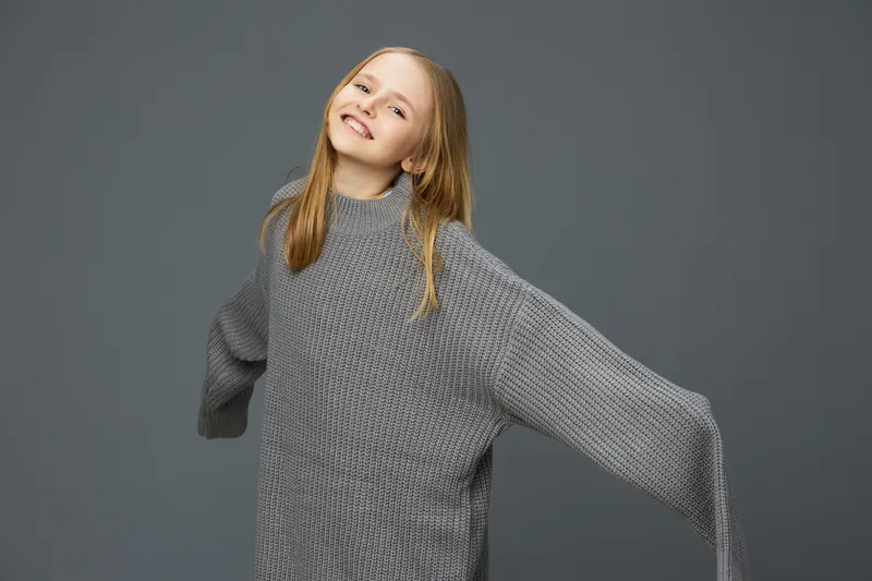 Happy girl in oversized gray sweater smiling against a neutral gray background, showcasing cozy winter style and a cheerful, playful emotion