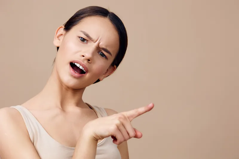 Expressive young woman showing disapproval with a raised finger, wearing a beige top against a neutral background, conveying strong emotions