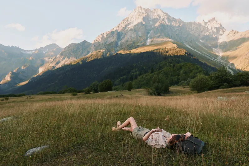 A young woman with long hair lies on her back in a grassy meadow surrounded by mountains, enjoying a peaceful moment in nature during a sunny day.