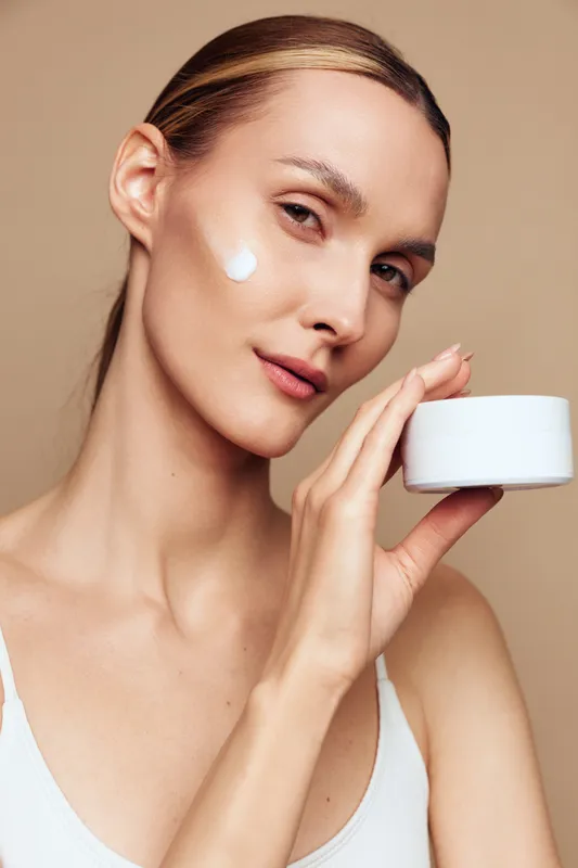 A young woman with clear skin applies white cream on her cheek while holding a skincare jar. The beige background highlights natural beauty and daily facial care routine.