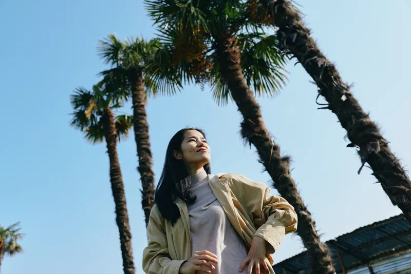 Woman posing outdoors among tall palm trees under a clear blue sky, wearing a light jacket and casual outfit, exuding confidence and a relaxed mood, sunny day, travel vibe, warm light and carefree