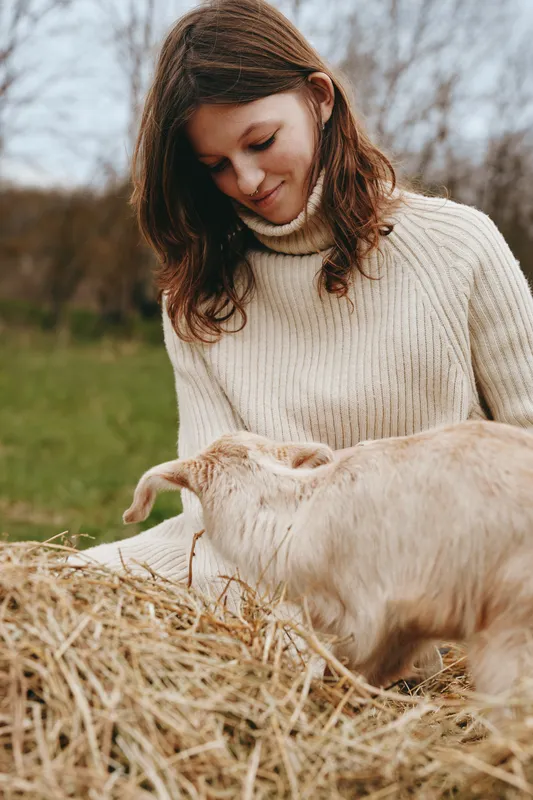 Calm woman in a cream knit sweater interacts with a friendly goat kid outdoors on a rural farm, capturing a heartwarming lifestyle moment amid hay and grass. The scene radiates warmth and gentle